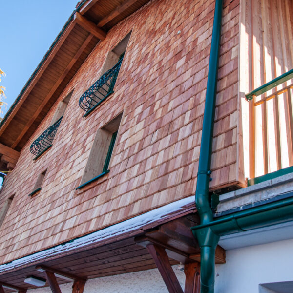 Holzfassade eines Wohnhauses mit grünem Balkon und geschwungenen Metallfenstergittern, fotografiert aus der Froschperspektive