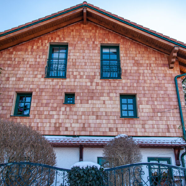 Frontalansicht eines Hauses mit neuer Holzschindelfassade, grün gerahmten Fenstern und Wintervegetation