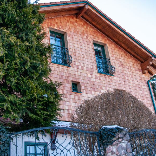 Schrägaufnahme eines Hauses mit Holzschindelfassade und Tannenbaum im Vordergrund